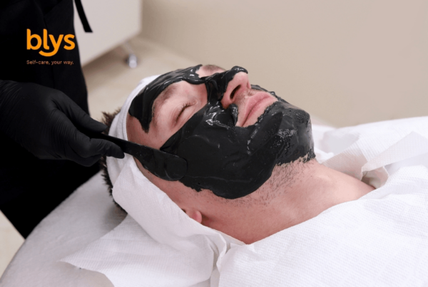 Male client relaxes during a facial treatment as an esthetician applies a glossy black mask, with the blys logo visible in the corner.