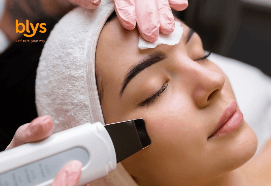 Woman relaxes during a facial cleansing treatment as a gloved esthetician uses an ultrasonic skin spatula on her cheek; soothing spa setting.