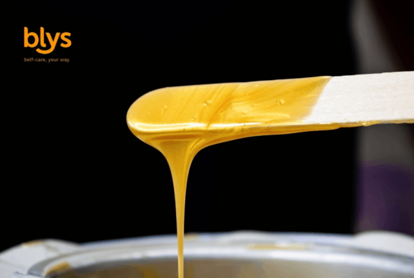 Close-up of golden skincare wax dripping from a spatula into a metal bowl against a black background; warm, glossy, professional tone.