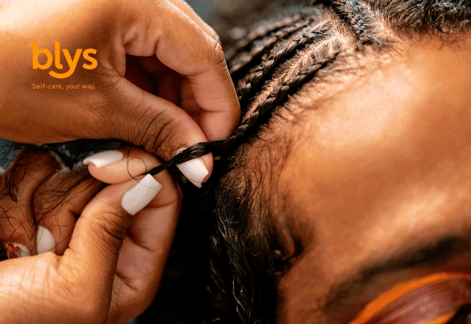 Close-up of hands braiding a Black person’s hair into neat cornrows, with white manicured nails and orange blys branding; warm, intimate beauty scene.