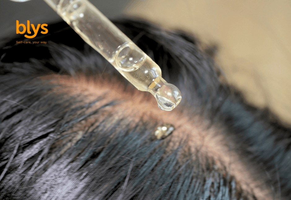 Close-up of a dropper releasing clear serum onto an exposed scalp between dark hair strands, suggesting a focused, soothing scalp treatment.