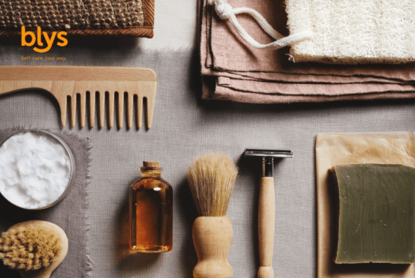 Flat lay of natural grooming essentials: wooden comb, shaving brush and razor, amber oil bottle, soap, towels, and exfoliating brush.
