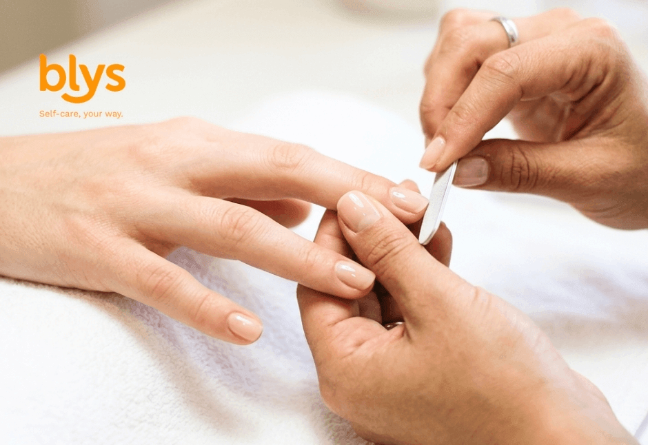 Close-up of a professional manicure: a nail technician gently shapes a client’s fingernail with a file. “blys” logo suggests self-care and relaxation.