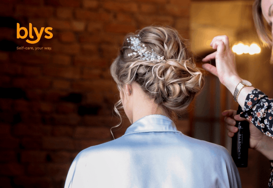Bride with an elegant curled updo and jeweled hairpiece sits as a stylist applies hairspray, creating a polished wedding look in warm light.