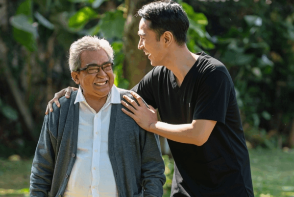 A younger man in a black shirt rests a hand on an older man's shoulder as they smile together in a leafy park, conveying warmth and affection.