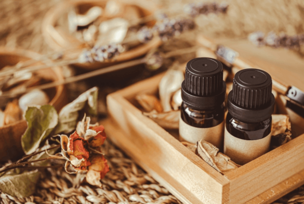 A warm, rustic arrangement of two dark essential-oil bottles in a wooden tray, surrounded by dried flowers, herbs, and natural aromatherapy massage scents.