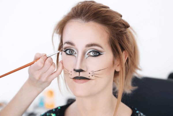 A makeup artist paints whiskers beside a woman’s nose; cat-style eyeliner, a black nose, and drawn whiskers complete her playful cat look.