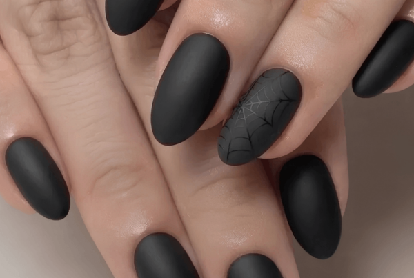 Close-up of manicured hands with matte black nails and a spiderweb accent nail, creating a sleek, moody Halloween look.