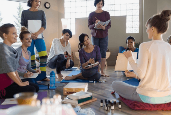 A diverse group of women coworkers sit in a sunlit studio, listening to a yoga practitioner with a clipboard; art supplies and notebooks suggest a warm, corporate retreat.