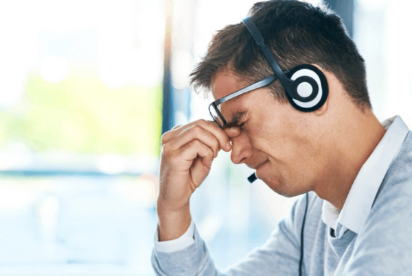 A male customer service worker wearing a headset and glasses pinches the bridge of his nose, facing burnout and stress in a bright office.