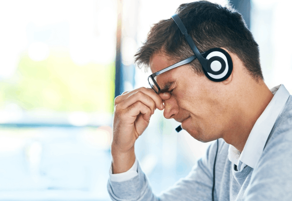 A male customer service worker wearing a headset and glasses pinches the bridge of his nose, facing burnout and stress in a bright office.