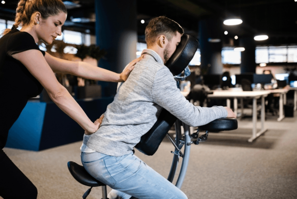 A massage therapist presses a man's back on a massage chair in a corporate setting, helping him relax during a focused, supportive chair massage session.