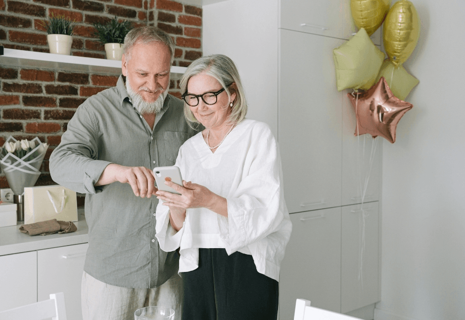Grandparents smile as they look at a smartphone in a bright kitchen, with gold and rose-gold balloons adding a warm, celebratory feel.