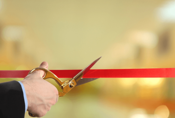 A hand in a dark suit uses gold-handled scissors to cut a red ribbon, marking a grand opening against a softly glowing gold background.