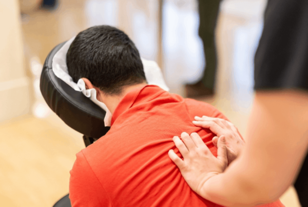 A person in a red shirt leans forward in a chair massage as a therapist gently presses their shoulders, creating a calm, relaxing wellness scene.