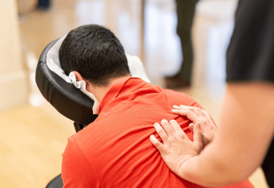 A person in a red shirt leans forward in a chair massage as a therapist gently presses their shoulders, creating a calm, relaxing wellness scene.