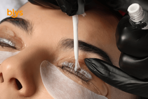Close-up of a woman receiving an eyelash tint treatment as a gloved technician applies product to her lashes above an under-eye patch; calm beauty-care scene.