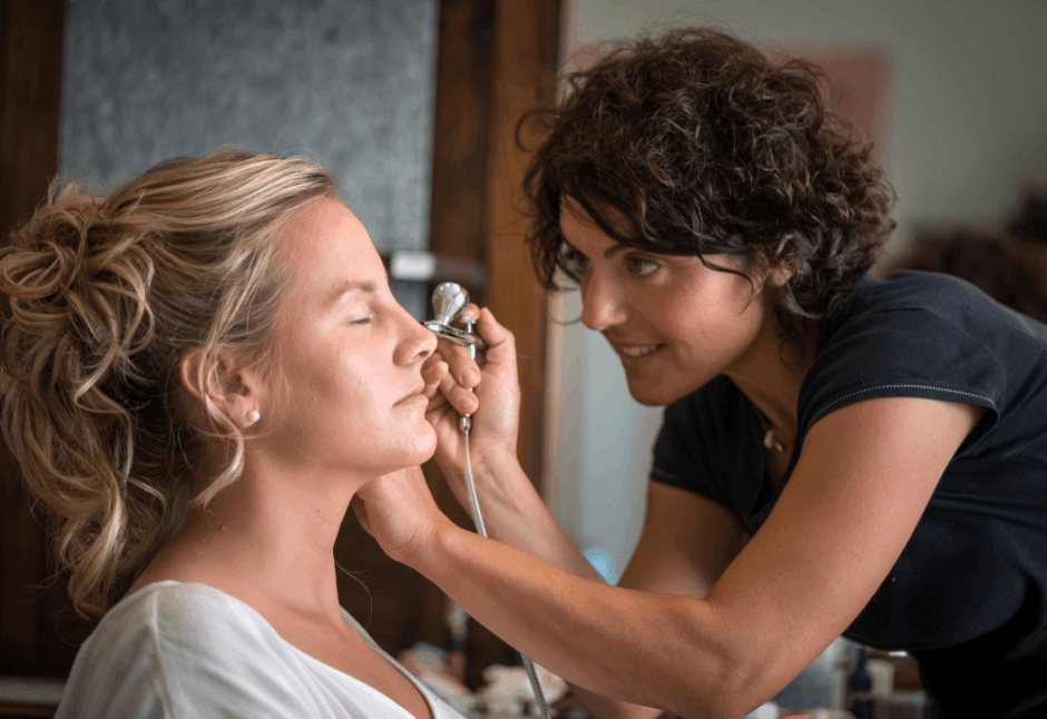 A makeup artist gently applies airbrush makeup to a seated blonde woman, who keeps her eyes closed; their close interaction feels calm and focused.