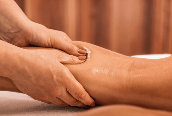 A therapist gently massages an oil-sheened client’s lower lipoedema legs on a treatment table, with warm curtains behind, creating a calm atmosphere.