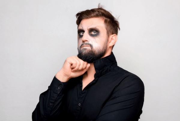 Bearded man in a black shirt poses against a plain gray background with dark, smudged eye makeup, chin resting on his fist and a gothic Halloween look.