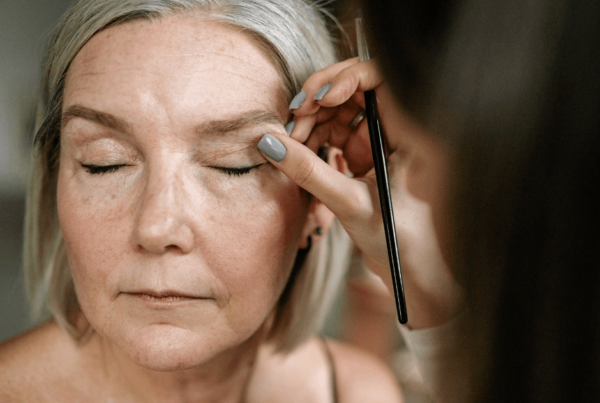 A makeup artist applies eyeliner to a silver-haired older woman with mature skin, creating a calm, intimate behind-the-scenes beauty moment.