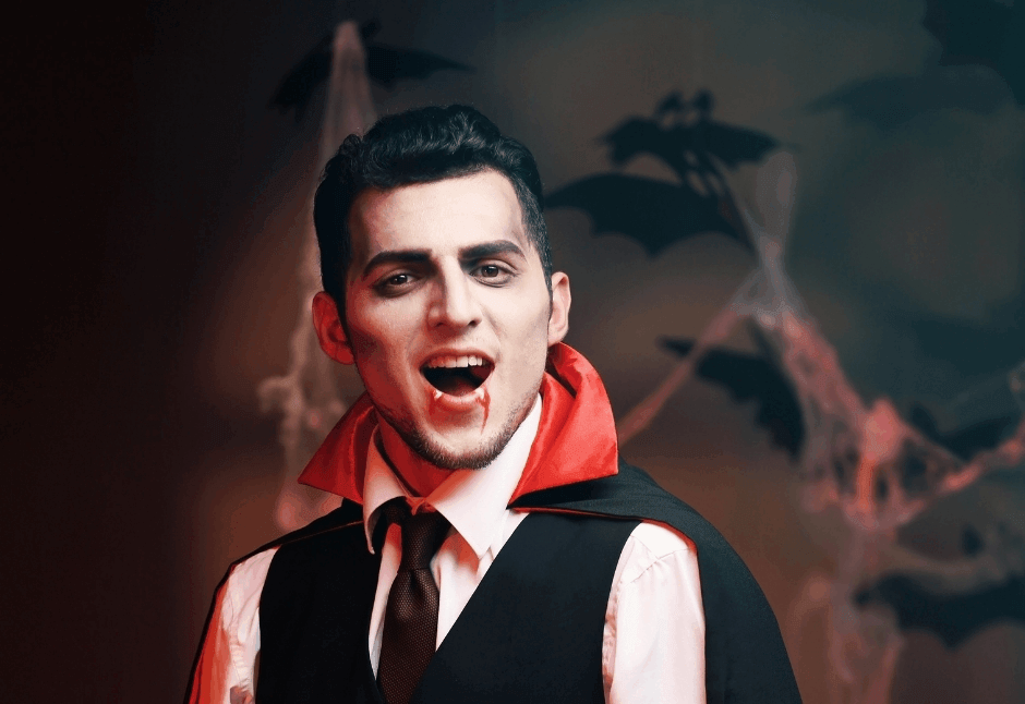 A young man in a vampire makeup, wearing a black vest and red-lined cape, bares fangs before a shadowy backdrop of bats and mist, looking theatrical.