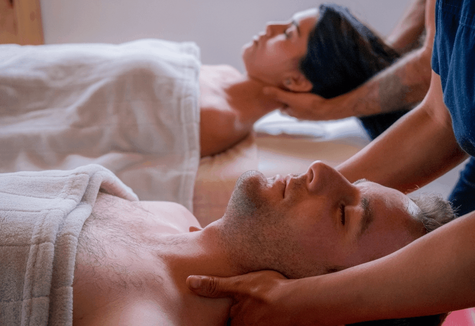 A couple relaxes under white towels as massage therapists gently work their necks and shoulders in a softly lit room, conveying calm and comfort.