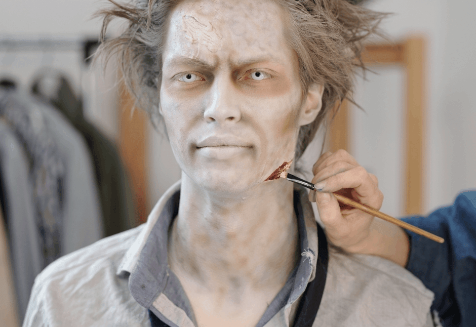 A pale-faced person with messy hair sits in costume as an unseen hand applies a bloody scratch to his neck, creating a tense zombie makeup scene.