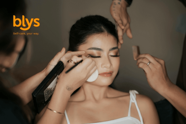 A makeup artist applies eyeliner to a serene woman while stylists adjust her hair; blys logo visible.