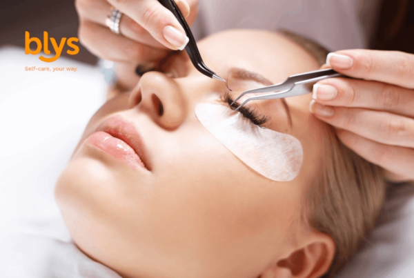 Close-up of a relaxed woman receiving lash extensions, with tweezers placing lashes above under-eye pads during a beauty treatment.