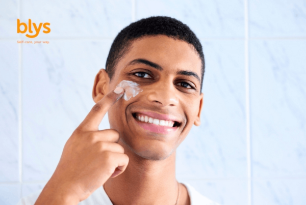 Smiling young man applies facial cream to his cheek in a bright tiled bathroom, conveying a fresh, confident skincare routine.