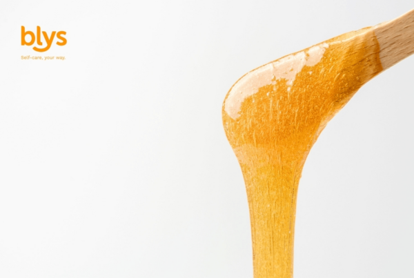 Close-up of a wooden spoon coated in sugar waxing for hair removal, stretching downward against a clean white background; blys logo appears at upper left.