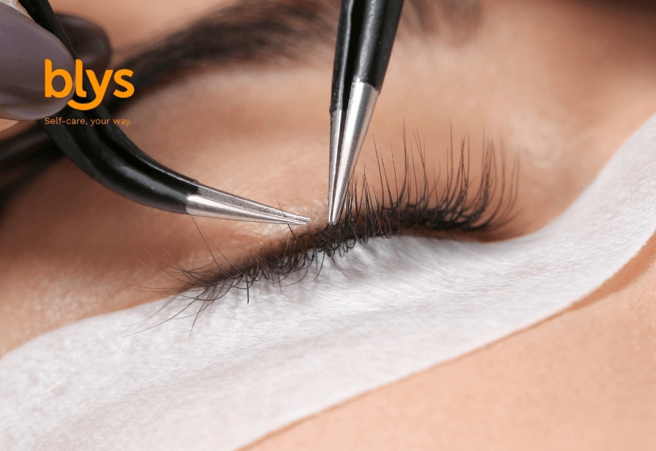 Close-up of a closed eye during eyelash extension treatment, with tweezers isolating lashes above a white under-eye pad; precise, calm beauty care.