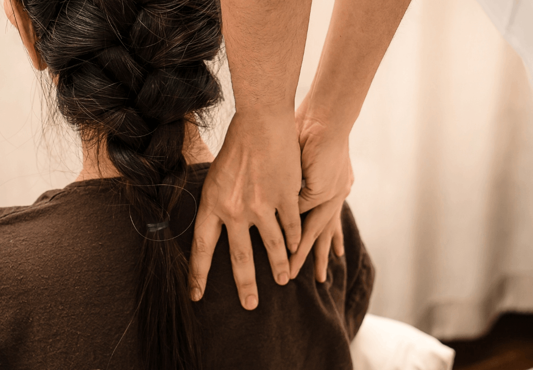 A Thai massage therapist’s hands gently massage a woman’s shoulders and upper back as she sits with braided hair, creating a calm, soothing wellness scene.