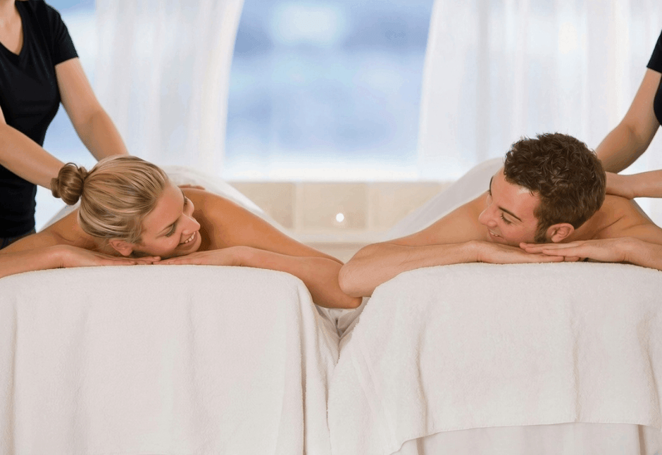 A relaxed couple enjoys a side-by-side mobile massage on white-covered tables, smiling at each other as therapists work in a bright, serene room.