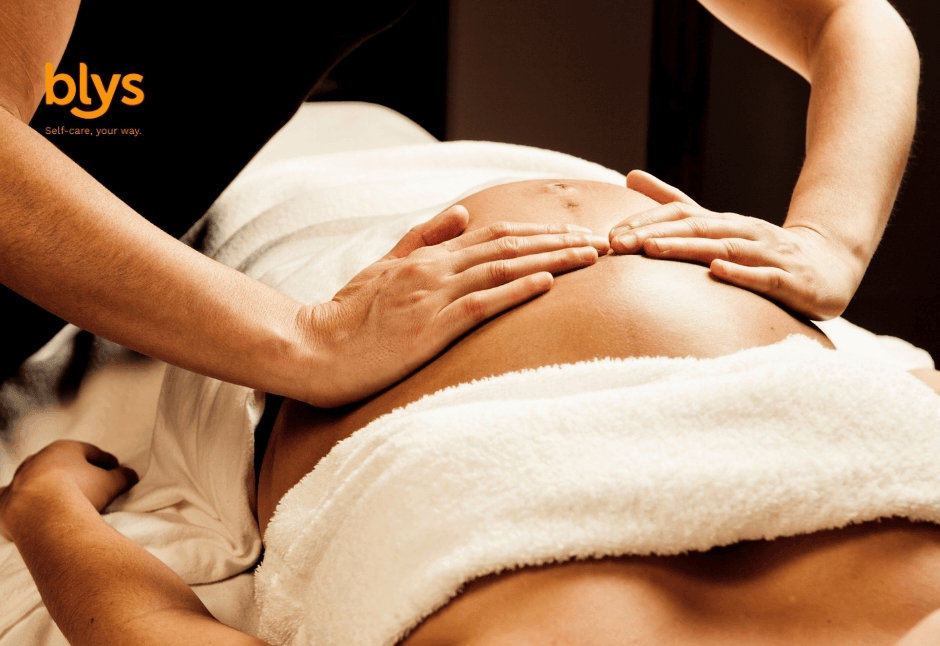 A therapist gently massages a pregnant person’s exposed belly in a warm setting, with white towels and soft lighting creating a calm, nurturing mood.