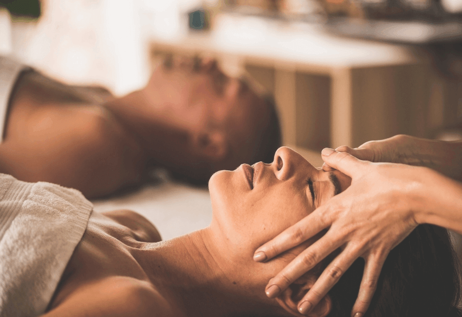 A relaxed couple receives a gentle head and face massage at a room, with a therapist’s hands at a woman's temples and jaw; warm light creates a soothing, peaceful mood.