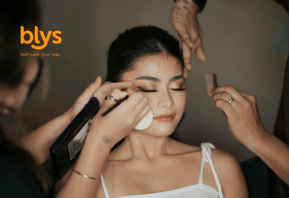A makeup artist applies eyeliner to a serene woman while stylists adjust her hair; blys logo visible.