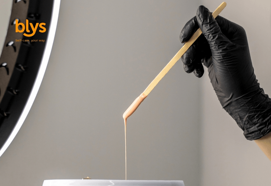 Gloved hand uses a wooden stick to drizzle wax into a container, with a ring light and orange “blys” self-care logo.