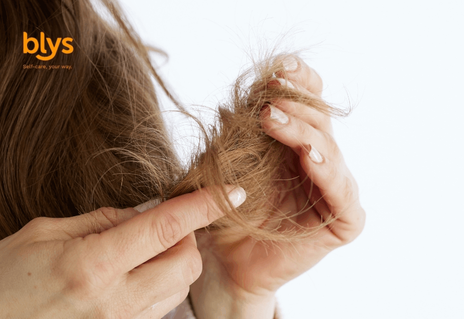 A close-up of a hand gently holding a lock of long brown hair with split ends against a light background, creating a calm, intimate beauty-care image. A small “blys” logo appears in the upper-left corner.