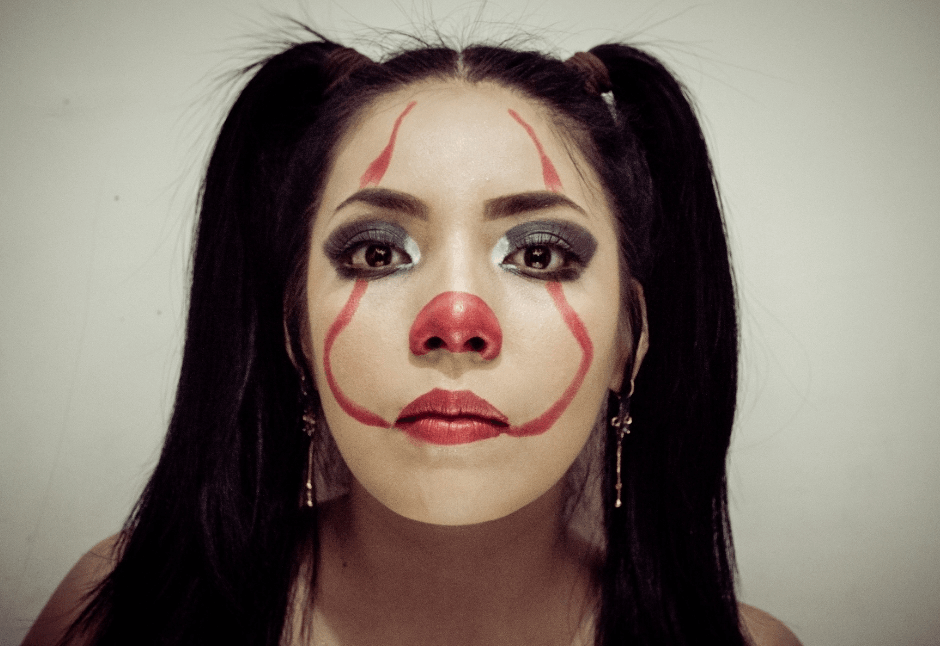 Close-up portrait of a woman with black pigtails and clown makeup; her neutral gaze feels somber and unsettling.
