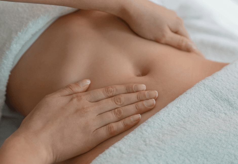 Close-up of a rounded bare abdomen gently cradled by two hands against a soft white towel during a post-lipo lymphatic drainage massage for fibrosis, conveying calm, tenderness, and self-care.