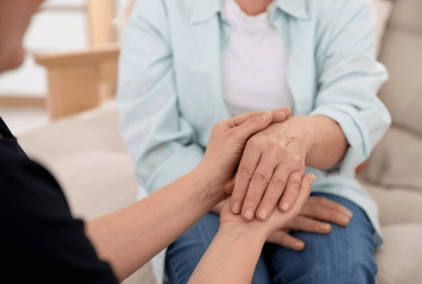 A manicure specialist gently holds an older woman’s hands in a bright room, offering comfort and support in a calm, compassionate moment.