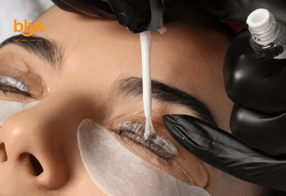 Close-up of a woman receiving an eyelash tint treatment as a gloved technician applies product to her lashes above an under-eye patch; calm beauty-care scene.
