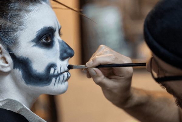 A makeup artist paints skeletal teeth on a person’s cheek, completing dramatic blue-and-white skeleton makeup in a focused backstage scene.