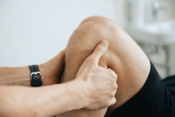 Close-up of a person’s hands gently massaging a bent, bare knee, with a black watch on one wrist; the intimate scene suggests knee swelling and stiffness after knee replacement.