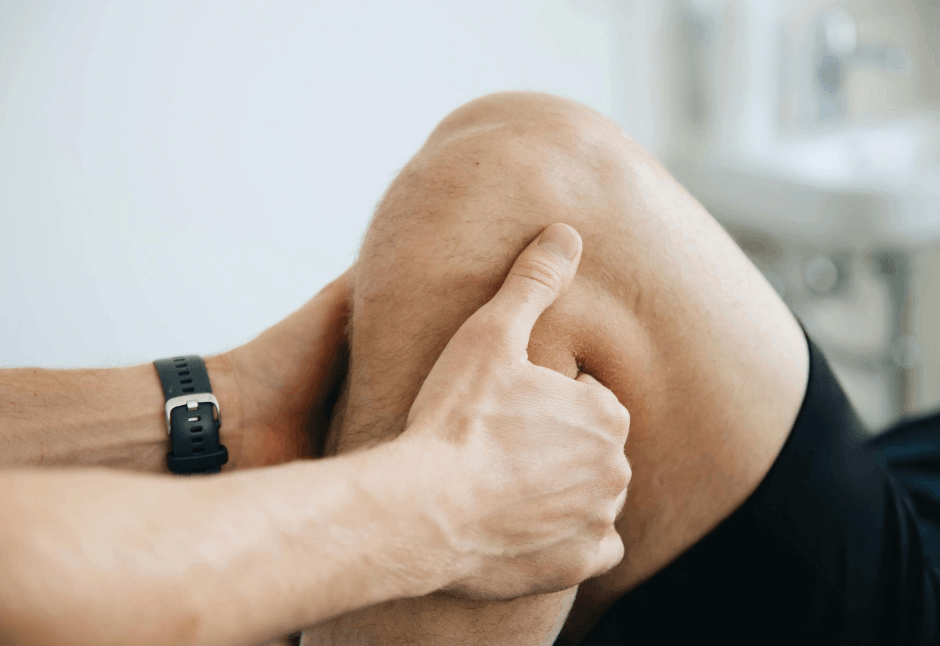 Close-up of a person’s hands gently massaging a bent, bare knee, with a black watch on one wrist; the intimate scene suggests knee swelling and stiffness after knee replacement.