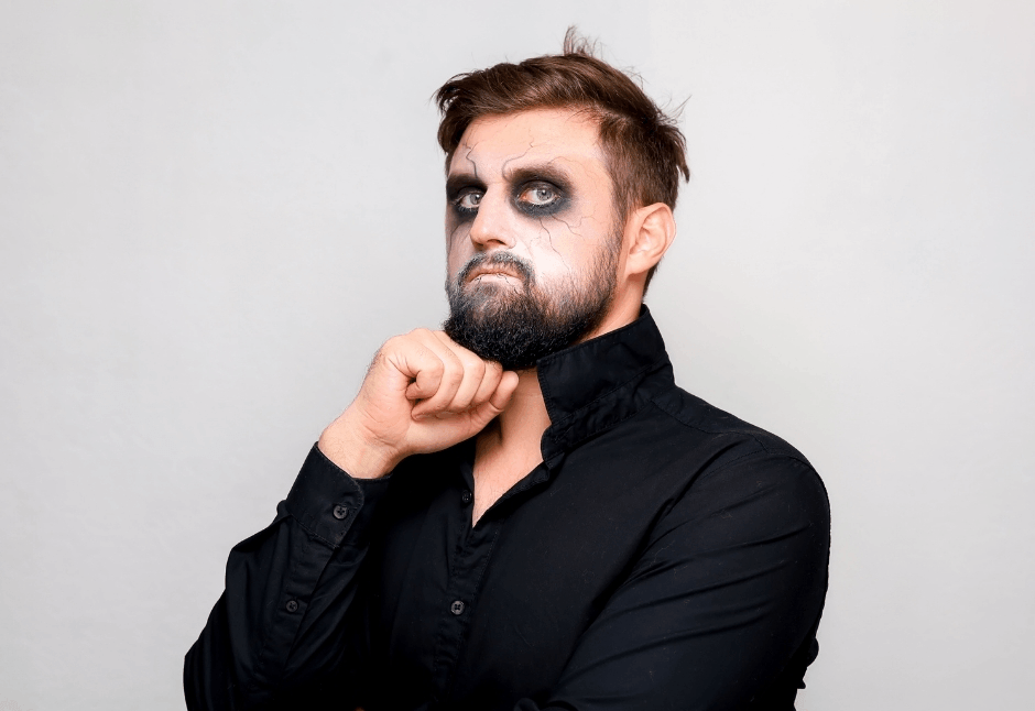 Bearded man in a black shirt poses against a plain gray background with dark, smudged eye makeup, chin resting on his fist and a gothic Halloween look.