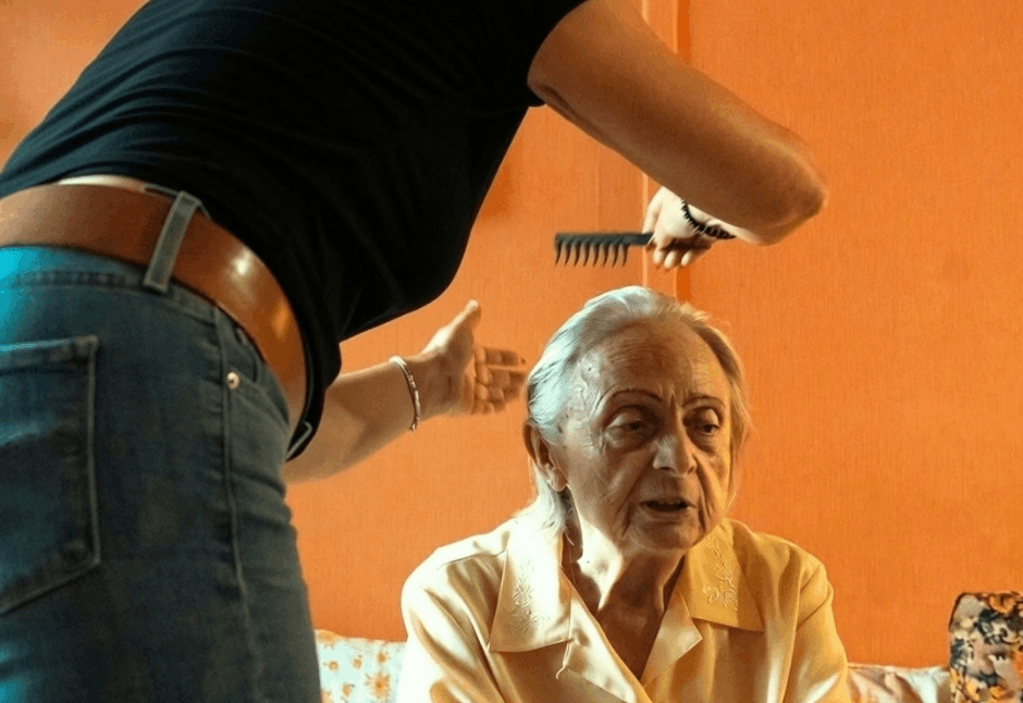 A stylist combs the hair of an elderly woman in a pale yellow blouse, seated against an orange wall; the intimate scene reflects homecare and feels warm and caring.