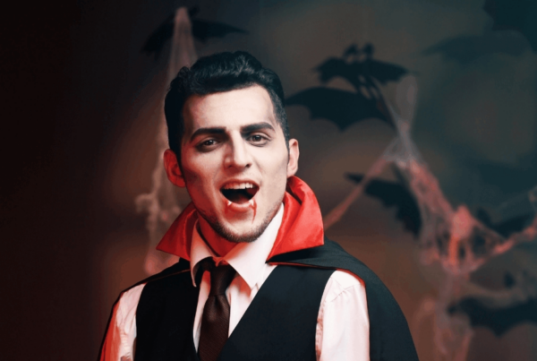 A young man in a vampire makeup, wearing a black vest and red-lined cape, bares fangs before a shadowy backdrop of bats and mist, looking theatrical.